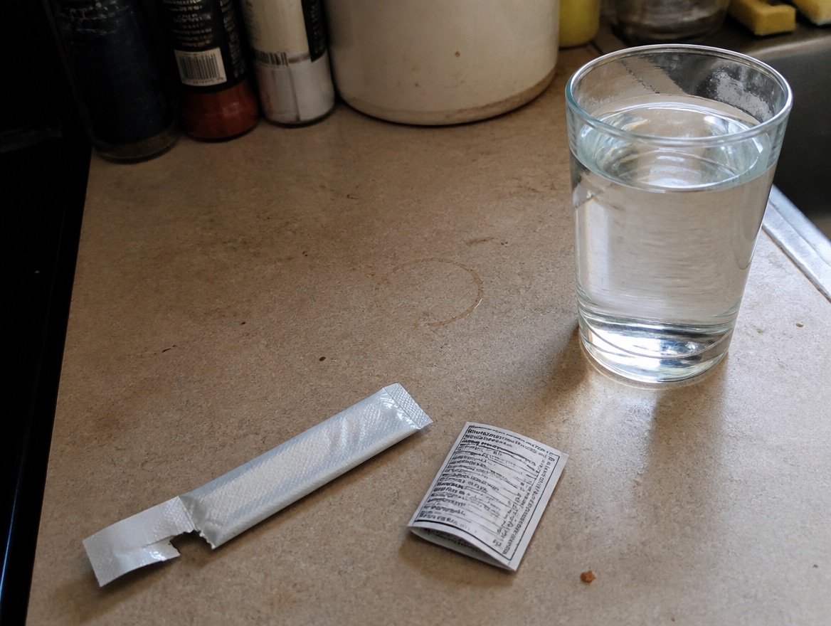 A torn electrolyte drink-mix stick pack lies flat on a kitchen counter beside a clear glass of water and a back-of-pack nutrition panel, available daylight from a side window