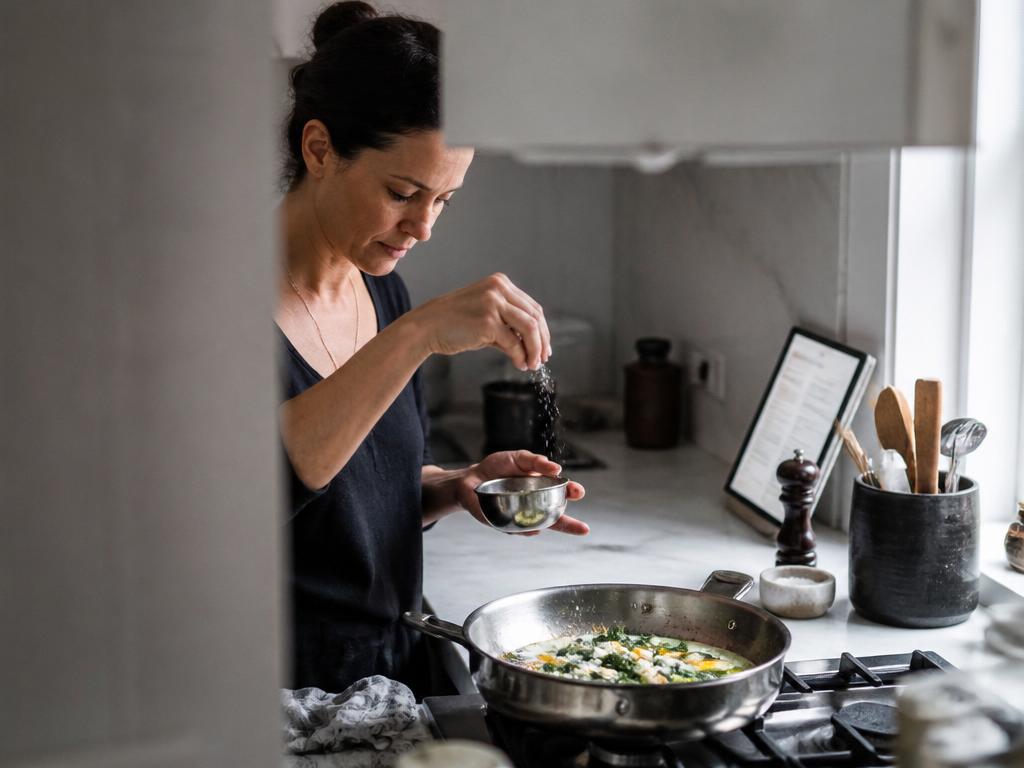 A woman sprinkles coarse salt from a height over a pan of eggs and greens cooking on a cool grey stovetop.