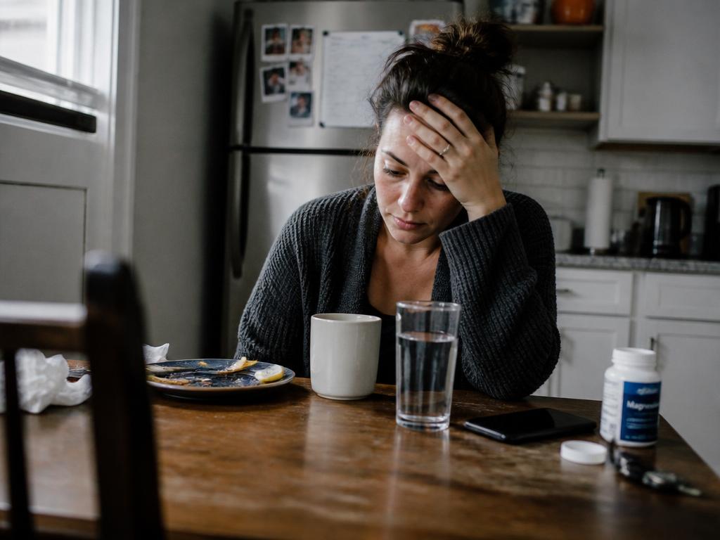 A woman in the first sluggish week of low-carb eating rubs her forehead at a kitchen table beside salted water and broth.